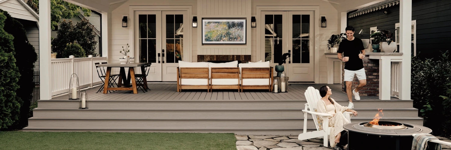 Couple on a grey composite deck with outdoor sofa and dining set; man carries mugs down steps toward a woman by a round fire pit—Fiberon Cottage decking.