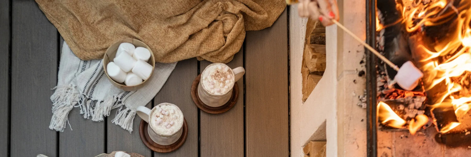 Cozy scene on grey Trex Biscayne composite decking beside an outdoor fire pit—mugs of hot chocolate, blanket, bowl of marshmallows, and a marshmallow roasting on a skewer.