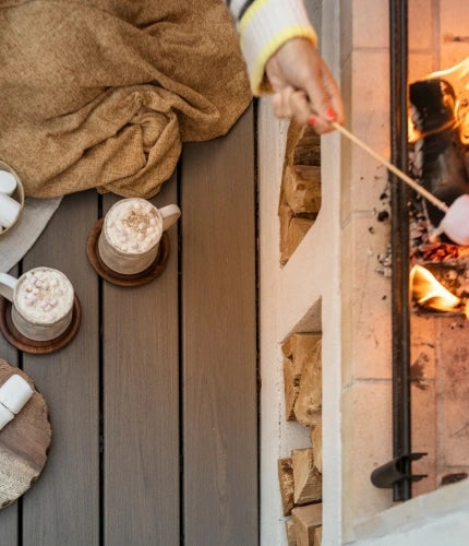 Cozy scene on grey Trex Biscayne composite decking beside an outdoor fire pit—mugs of hot chocolate, blanket, bowl of marshmallows, and a marshmallow roasting on a skewer.